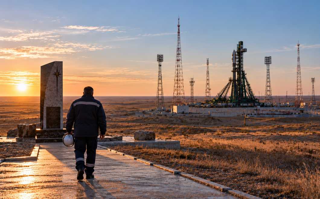 Работник с белой каской идёт по дорожке к памятнику на фоне стартового комплекса