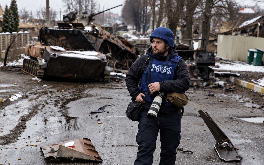 Фотожурналист в синей каске и жилете PRESS стоит с профессиональной камерой на разрушенной городской улице